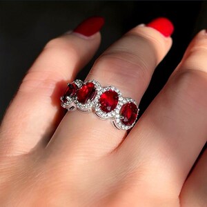 Könnte beinhalten: Ein silberner Ring mit fünf ovalen, tiefroten Edelsteinen, die jeweils von kleineren, klaren Steinen umgeben sind. Der Ring befindet sich an einem Finger mit rotem Nagellack.
