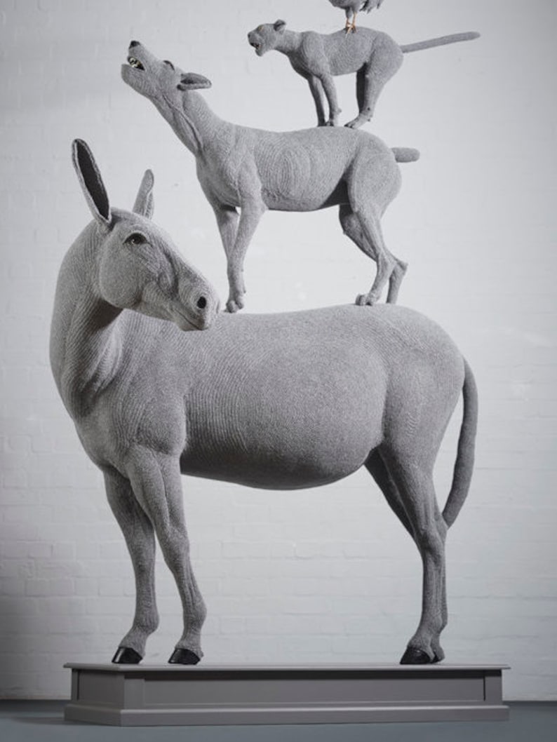 May include: A gray sculpture of a donkey, a wolf, a lion, and a bird stacked on top of each other. The donkey is at the base, with the wolf standing on its back. The lion is on the wolf, and the bird is on the lion.