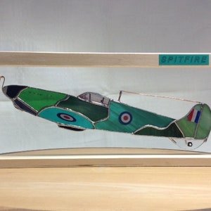 May include: A stained glass model of a Spitfire aircraft, displayed in a wooden frame. The plane is predominantly green and teal, with roundels and a French flag detail. The word "SPITFIRE" is printed on a green label on the frame.