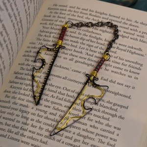May include: Two sword-shaped bookmarks in black and gold wire, with red and gold detailing on the handles, linked by a chain, lying on the open pages of a book.