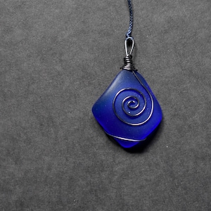 Peut inclure: Un pendentif en verre bleu avec un motif en spirale de fil argenté. Le pendentif est attaché à un cordon noir.