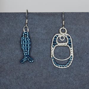 Könnte beinhalten: Zwei handgefertigte Ohrringe. Einer hat die Form eines Fisches, der andere die Form eines Getränkedosen-Tabs. Beide Ohrringe sind aus Silberdraht und blauen Perlen gefertigt. Der Fisch-Ohrring ist etwa 2,5 cm lang.