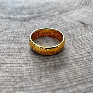 May include: A gold ring with engraved text and decorative patterns. The ring is set against a textured, grey wooden surface. The ring's design features intricate details and a polished finish.