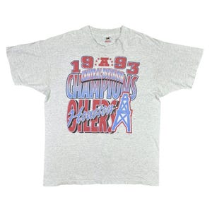 May include: A gray t-shirt with a vintage design featuring the text "1993 Central Division Champions Houston Oilers" in red, blue, and white. The design also includes a stylized oil derrick.