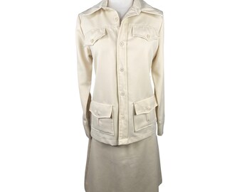 May include: A cream-colored two-piece outfit with a long-sleeved collared jacket and a matching skirt. The jacket has a button-up front, two flap pockets on the chest, and two large flap pockets on the bottom. The skirt is knee-length and has a straight silhouette.
