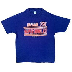 May include: A vintage blue t-shirt with the Chicago Bears logo and the text "Super Bowl XX" in orange and red. The text "January 26, 1986 New Orleans" is printed below the logo.