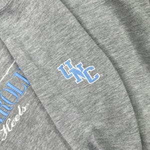 May include: Close-up of a grey sweatshirt sleeve with the text "ROLL Heels" in white and a light blue "UNC" logo. The sleeve is made of a soft, textured fabric.