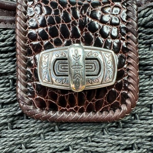 Puede incluir: Primer plano de un bolso de cuero marr&oacute;n con un cierre plateado. El cierre tiene un dise&ntilde;o decorativo con un motivo floral.