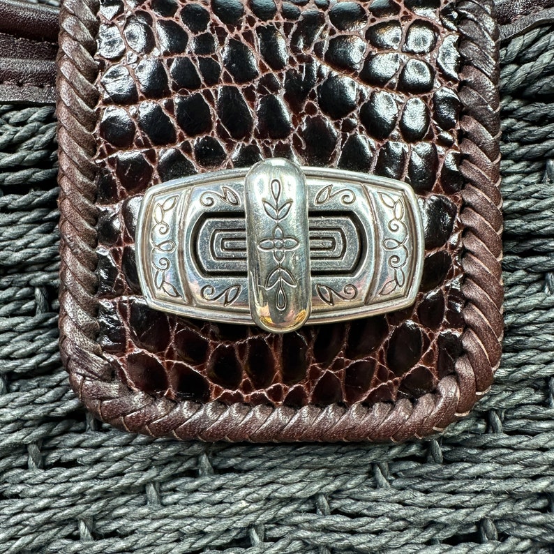 Puede incluir: Primer plano de un bolso de cuero marr&oacute;n con un cierre plateado. El cierre tiene un dise&ntilde;o decorativo con un motivo floral.