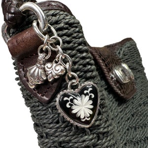 Puede incluir: Primer plano de un bolso tejido de color gris oscuro con una correa de cuero marr&oacute;n y un dije de plata con un colgante en forma de coraz&oacute;n. El colgante tiene un fondo de esmalte negro con un dise&ntilde;o floral blanco.