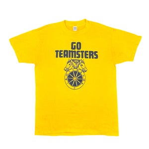 May include: Yellow t-shirt with the text "GO TEAMSTERS" in dark blue. Below the text is a graphic of three horses above a wheel with text around the perimeter. A classic crew neck tee.
