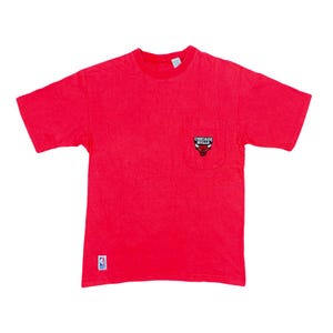 May include: Red t-shirt with a pocket featuring the Chicago Bulls logo. The shirt has a crew neck and short sleeves. A small NBA logo is visible near the bottom. The shirt is a solid red color.