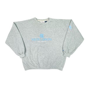 May include: A grey sweatshirt featuring the text "North Carolina Tar Heels" in light blue. The long-sleeved, crew-neck top has a light blue logo on the left sleeve. The sweatshirt is a casual style.