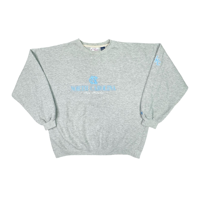 May include: A grey sweatshirt featuring the text "North Carolina Tar Heels" in light blue. The long-sleeved, crew-neck top has a light blue logo on the left sleeve. The sweatshirt is a casual style.