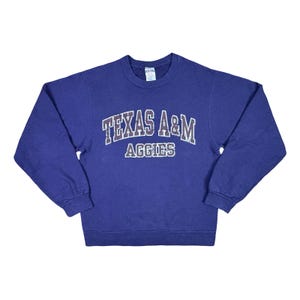 May include: A blue crewneck sweatshirt with the words "TEXAS A&M AGGIES" in white and maroon lettering. The sweatshirt is a solid blue color and has long sleeves.