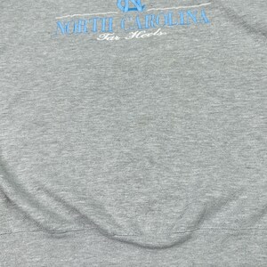 May include: Grey sweatshirt featuring the text "NORTH CAROLINA" in light blue, arched across the top, and "Tar Heels" below. The fabric has a soft, textured appearance.