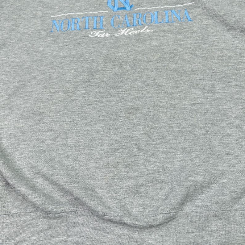 May include: Grey sweatshirt featuring the text "NORTH CAROLINA" in light blue, arched across the top, and "Tar Heels" below. The fabric has a soft, textured appearance.