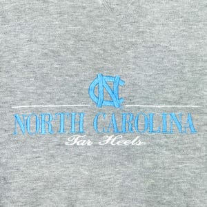 May include: A grey sweatshirt featuring light blue embroidered text reading "NORTH CAROLINA" with a matching logo above. The words "Tar Heels" are embroidered in white below.