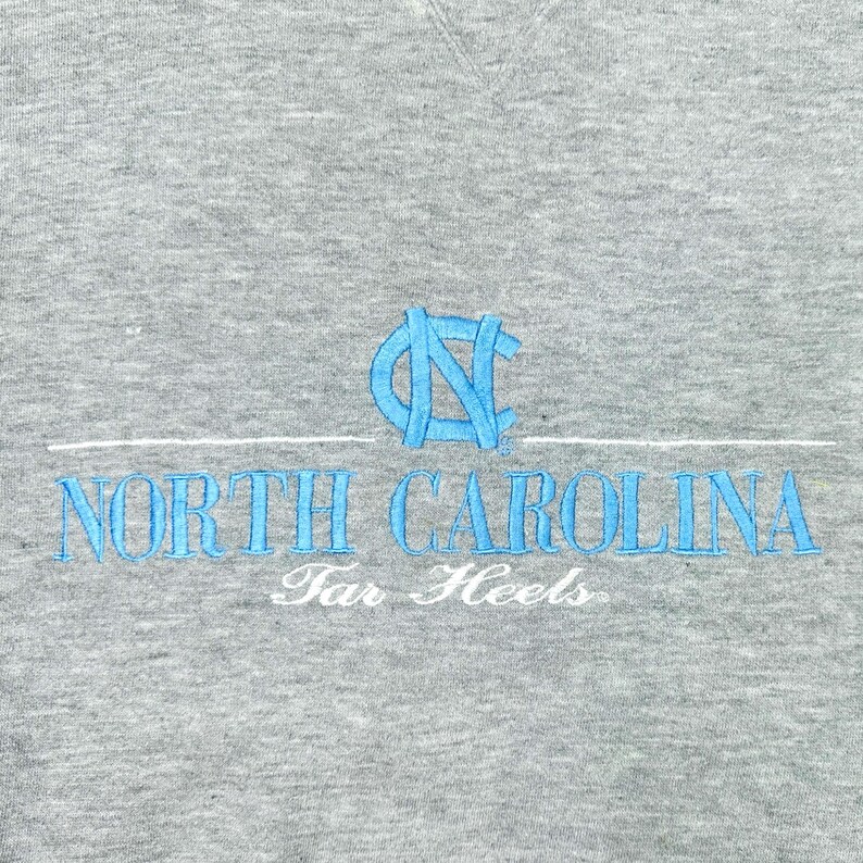 May include: A grey sweatshirt featuring light blue embroidered text reading "NORTH CAROLINA" with a matching logo above. The words "Tar Heels" are embroidered in white below.
