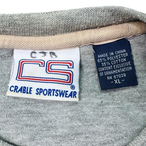 May include: A close-up of a grey Crable Sportswear shirt label. The label shows the letters "CS" in a red and blue design. A smaller label indicates the shirt is made in China, with 65% polyester and 35% cotton.