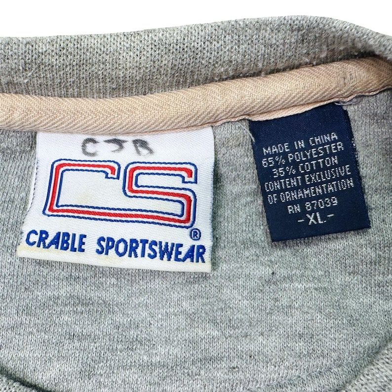 May include: A close-up of a grey Crable Sportswear shirt label. The label shows the letters "CS" in a red and blue design. A smaller label indicates the shirt is made in China, with 65% polyester and 35% cotton.