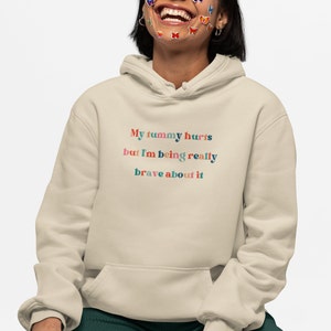 Puede incluir: Una sudadera con capucha beige con el texto "My tummy hurts but I'm being really brave about it" en letras de colores.