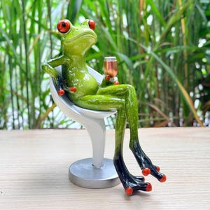 Può includere: Una statuetta di rana verde con grandi occhi rossi e punte delle dita arancioni, seduta su una sedia moderna bianca. La rana tiene in mano un piccolo calice color rame, con le lunghe zampe che si estendono verso il basso.