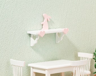 May include: White shelf with pink heart accents and a pink bunny figurine. The shelf is mounted on a light green wall. There is a white table and two white chairs in the background.
