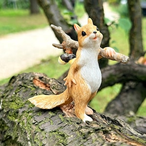 May include: A squirrel figurine with light brown fur and a white chest, standing upright while holding a small wooden branch over its shoulder. The branch features a small, round, dark-coloured bundle tied to it with a thin brown cord.