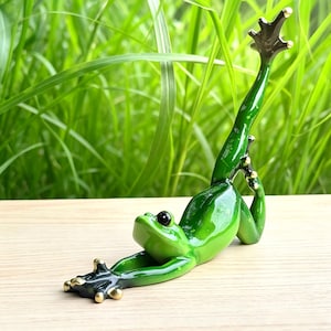 May include: A glossy green frog figurine in a stretching pose with one leg extended upwards. The frog has a smooth, vibrant green body, dark black eyes, and gold-coloured accents on the tips of its toes and fingers, creating a detailed appearance.