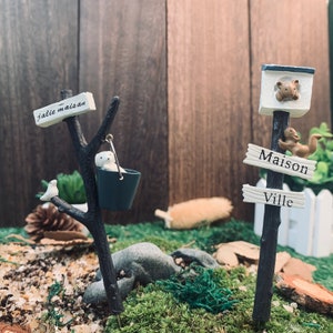 Könnte beinhalten: Zwei Miniatur-Holzschildchen mit weiß bemalten Schildern. Das Schild links zeigt "j'allie maison" und das Schild rechts zeigt "Maison" und "Ville". Die Schilder sind mit Miniaturtieren und einem Eimer verziert.