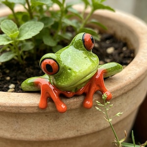 May include: A decorative ceramic frog figurine featuring a bright green body with a glossy finish, prominent bulging orange eyes with black pupils, and vibrant orange feet, positioned to appear as if it is clinging to the rim of a plant pot.