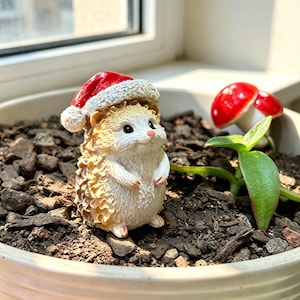 May include: A small, light-coloured hedgehog figurine wearing a red and white Santa hat. The hedgehog has a textured, spiky back, a smooth cream-coloured belly, and a small pink nose. It stands upright with its front paws tucked near its chest.