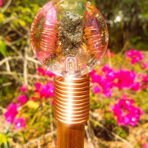 Pode incluir: Um topo de varinha transparente em forma de esfera com cristais embutidos, fio dourado e um centro escuro e texturizado. A varinha tem uma haste cor de cobre e um detalhe de fio enrolado perto da esfera. O fundo apresenta flores cor-de-rosa.