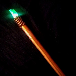 Pode incluir: Uma varinha de madeira com um cristal verde brilhante no topo. A varinha é marrom e tem uma superfície texturizada.