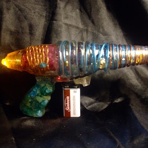 May include: A colorful, homemade toy ray gun with a clear plastic body, wrapped in copper and blue wire. The handle is made of green and blue resin with a 9V battery attached.