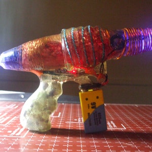 May include: A clear plastic toy gun with a red, orange, and blue design. The gun has a copper coil wrapped around the barrel and a 9V battery attached to the bottom.