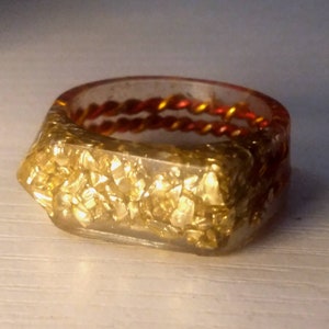 May include: A clear resin ring with gold flakes embedded in the resin. The ring has a red and gold braided cord wrapped around the band.