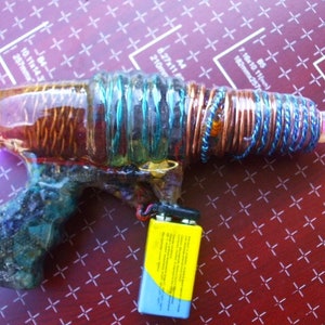 May include: A homemade toy gun made from a clear plastic bottle, wrapped with copper wire and blue string. The gun has a pink light at the end of the barrel and a yellow battery pack attached to the bottom.