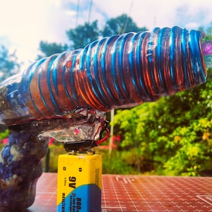 May include: A homemade water gun with a clear plastic barrel wrapped in blue and copper wire. The gun has a blue and black handle and is powered by a 9V battery.