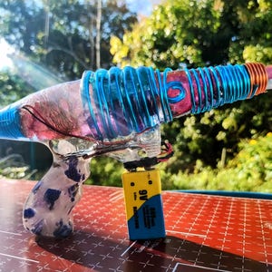 May include: A handmade ray gun with a clear body, blue and copper wire accents, and a 9-volt battery. The handle is white with blue and purple accents. The ray gun is set against a blurred background of green foliage.