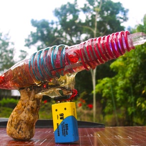 May include: A clear plastic toy gun with a red and blue coil wrapped around it. The gun has a wooden handle and a purple crystal at the end of the barrel. A 9V battery is attached to the side of the gun.