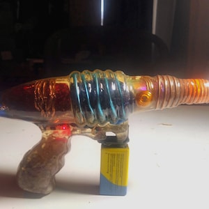 May include: A glass, multi-colored, futuristic-looking toy gun with a clear, brown, and orange barrel. The gun has a clear, brown, and blue handle. The gun is sitting on a white surface with a yellow battery pack.