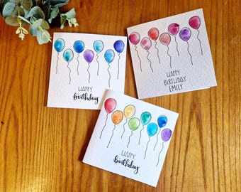 May include: Three watercolor Happy Birthday cards with colorful balloons. Each card features a different arrangement of balloons in shades of blue, teal, purple, red, orange, and yellow. The cards are on a wooden surface.