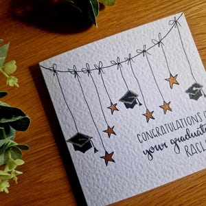 May include: A square white greeting card with a textured surface featuring a black ink illustration of a string with bows hanging down, decorated with three black graduation caps and five copper-coloured stars. The text reads: CONGRATULATIONS ON your graduation RACHEL.