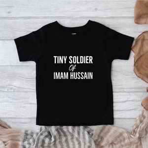 May include: Black short-sleeved t-shirt with white text that reads "Tiny Soldier of Imam Hussain".