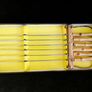 May include: A set of six metal skewers with bamboo handles in a yellow box. The skewers are arranged in a row, with the handles on the right side of the box.