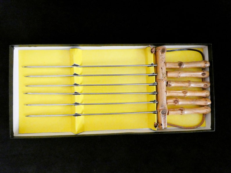 May include: A set of six metal skewers with bamboo handles in a yellow box. The skewers are arranged in a row, with the handles on the right side of the box.