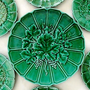 Peut inclure: Une collection d'assiettes circulaires en céramique verte présentant un motif en relief de feuilles de vigne et de grappes de raisin au centre. Chaque assiette possède un bord festonné et une finition brillante, d'un vert émeraude éclatant.