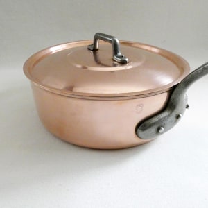 May include: A copper saucepan with a lid and a long, riveted handle. The pot has a shiny, reflective surface and a dark metal handle. The lid has a small, square handle. Kitchenware.